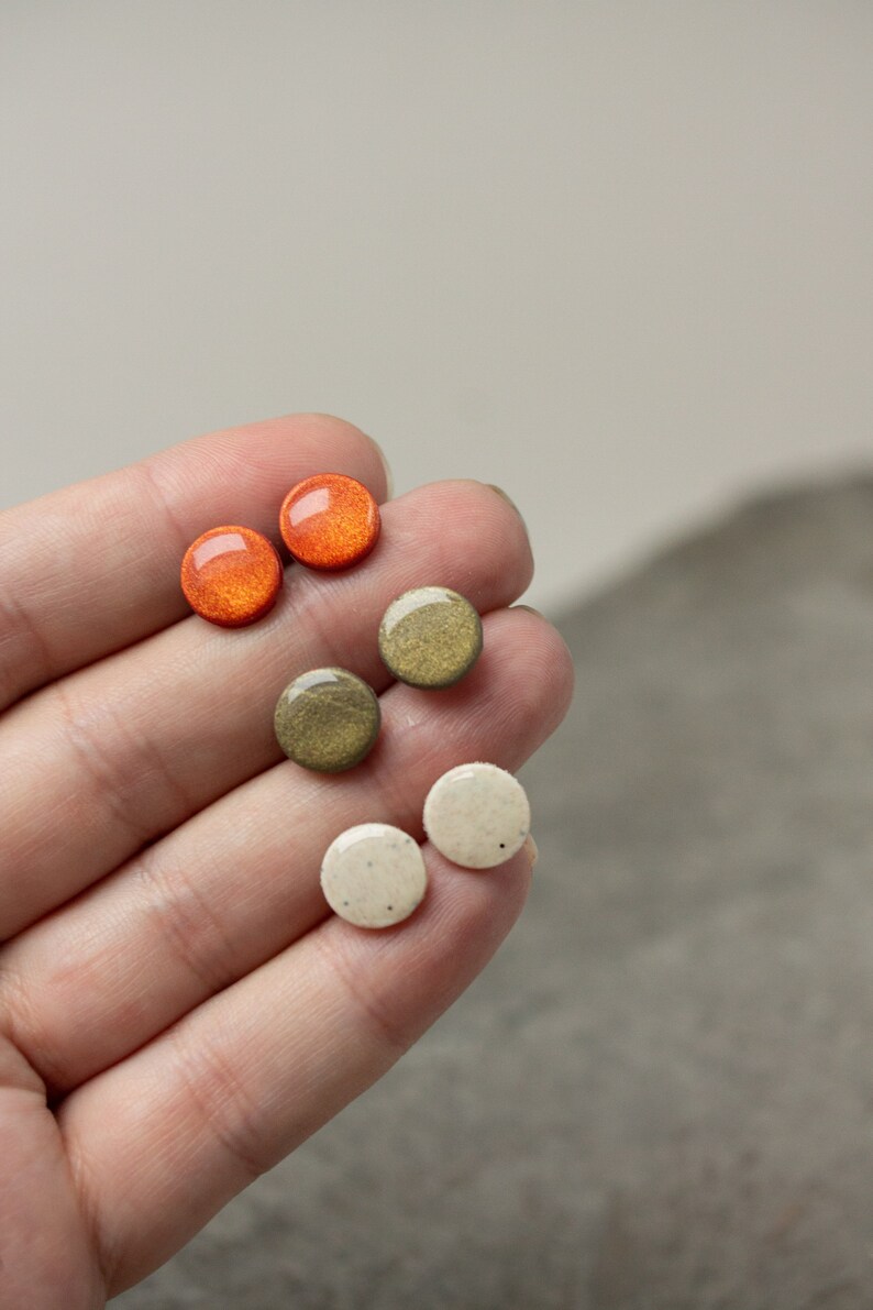 May include: Six small round ceramic earrings in three colors: orange, green, and white. The earrings are displayed on a hand.