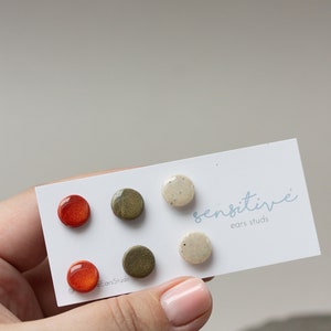 May include: Set of six ceramic earrings in three colors: orange, green, and white. The earrings are round and have a speckled finish. The card reads "sensitive ears studs".