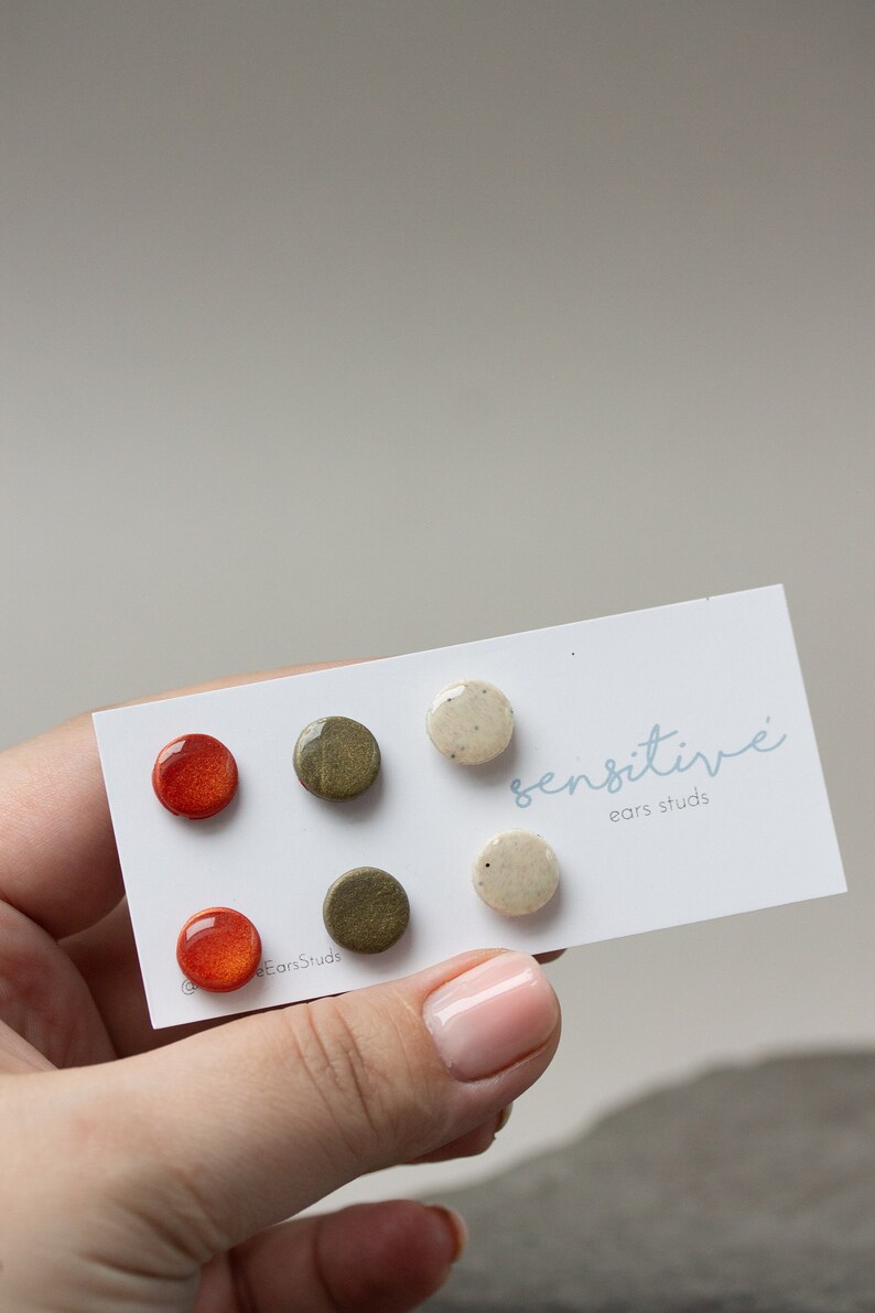 May include: Set of six ceramic earrings in three colors: orange, green, and white. The earrings are round and have a speckled finish. The card reads "sensitive ears studs".