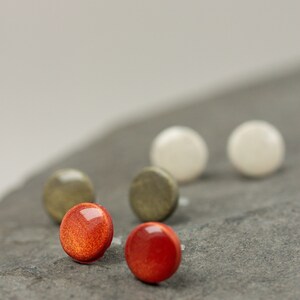 May include: A set of six round stud earrings in various colors, including red, white, and green. The earrings are displayed on a gray stone surface.