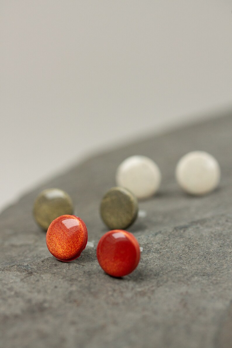 May include: A set of six round stud earrings in various colors, including red, white, and green. The earrings are displayed on a gray stone surface.