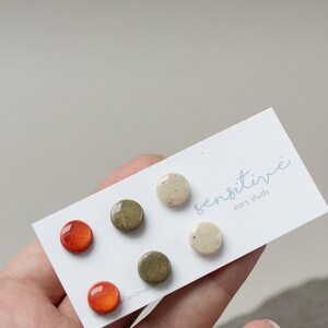 May include: Set of six small, round ceramic earrings in shades of orange, green, and white. The earrings are displayed on a white card with the text "sensitive ears studs".
