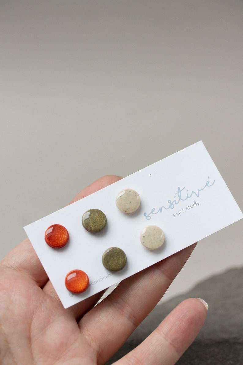 May include: Set of six small, round ceramic earrings in shades of orange, green, and white. The earrings are displayed on a white card with the text "sensitive ears studs".