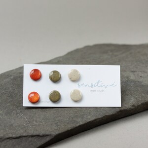 May include: Set of three round ceramic earrings in orange, green, and white. The earrings are displayed on a white card with the text "sensitive" and "ear studs".