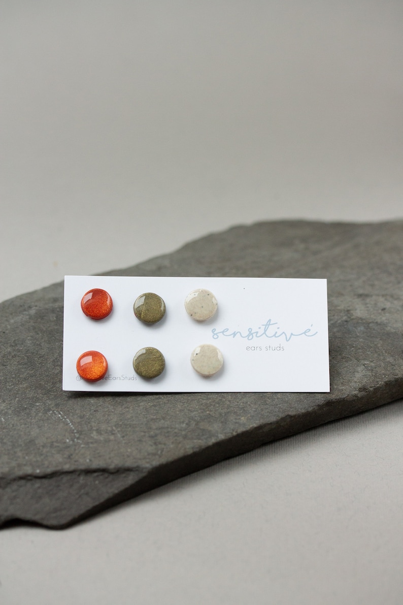 May include: Set of three round ceramic earrings in orange, green, and white. The earrings are displayed on a white card with the text "sensitive" and "ear studs".