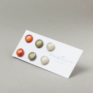 May include: Set of four small, round, stud earrings. Two are orange, one is green, and one is beige. The earrings are displayed on a white card with the text "sensitive ear studs".