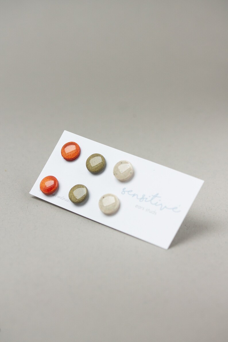 May include: Set of four small, round, stud earrings. Two are orange, one is green, and one is beige. The earrings are displayed on a white card with the text "sensitive ear studs".
