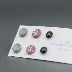May include: A set of six hypoallergenic earrings, including two pairs of gray, one pair of purple with gold accents, and one pair of black. The earrings are displayed on a white card with the text "sensitive ears studs" printed on it.