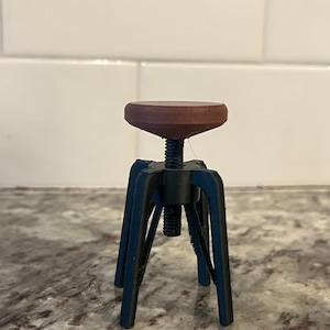 May include: A miniature adjustable stool featuring a round, brown seat and dark blue legs. The seat is supported by a black threaded rod and a base with four legs. The stool is designed for stability and is suitable for various uses.