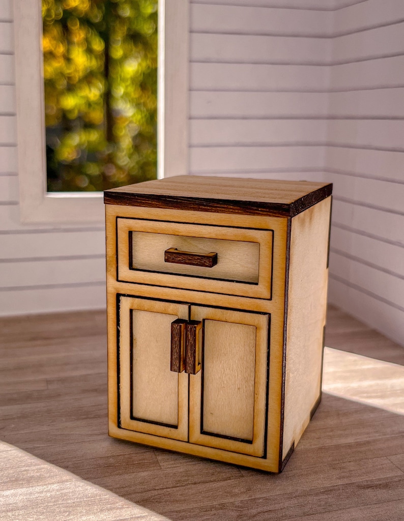 Puede incluir: Un peque&ntilde;o armario de madera con un solo caj&oacute;n y dos puertas. El armario es de color marr&oacute;n claro con un ribete marr&oacute;n m&aacute;s oscuro. Las puertas tienen un dise&ntilde;o sencillo con un solo tirador.