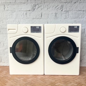Peut inclure: Deux machines à laver miniatures blanches avec des détails noirs. Chaque machine a une porte ronde à bord noir et un écran numérique. Les appareils sont placés contre un mur de briques blanches et un sol en bois clair.