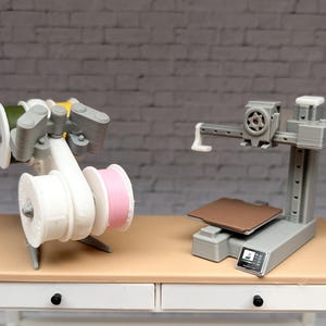 May include: Two miniature 3D printers are displayed on a small table. One is a white and grey filament holder with spools of green, yellow, and pink filament. The other is a grey printer with a brown print bed and a small screen.