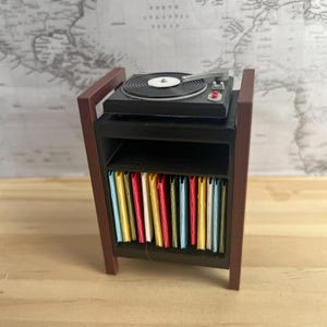Puede incluir: Miniatura de tocadiscos negro sobre una estantería negra con una colección de portadas de álbumes coloridas. La estantería tiene soportes de madera marrón y está colocada contra un mapa del mundo. Se ve un botón rojo en el tocadiscos.
