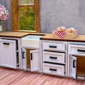 Puede incluir: Gabinetes de cocina en miniatura blancos con manijas negras y puertas abiertas. Los gabinetes tienen una encimera de madera y un peque&ntilde;o fregadero blanco. Hay tres pasteles en una caja de cuadros rojos y blancos sobre la encimera.