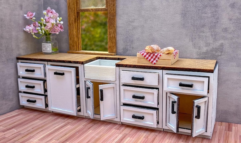 Puede incluir: Gabinetes de cocina en miniatura blancos con manijas negras y puertas abiertas. Los gabinetes tienen una encimera de madera y un peque&ntilde;o fregadero blanco. Hay tres pasteles en una caja de cuadros rojos y blancos sobre la encimera.