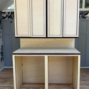 May include: A miniature wooden kitchen cabinet and countertop set. The upper cabinet has four doors, and the countertop has two open spaces. The wood is light-colored with dark brown trim, designed for a dollhouse or miniature setting.