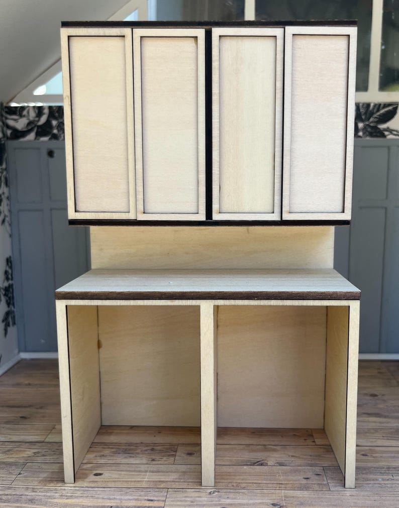 May include: A miniature wooden kitchen cabinet and countertop set. The upper cabinet has four doors, and the countertop has two open spaces. The wood is light-colored with dark brown trim, designed for a dollhouse or miniature setting.
