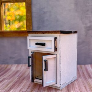 Puede incluir: Un armario blanco con una tapa de madera, un caj&oacute;n y dos puertas. Las puertas est&aacute;n abiertas, revelando un interior vac&iacute;o. El armario tiene tiradores negros.