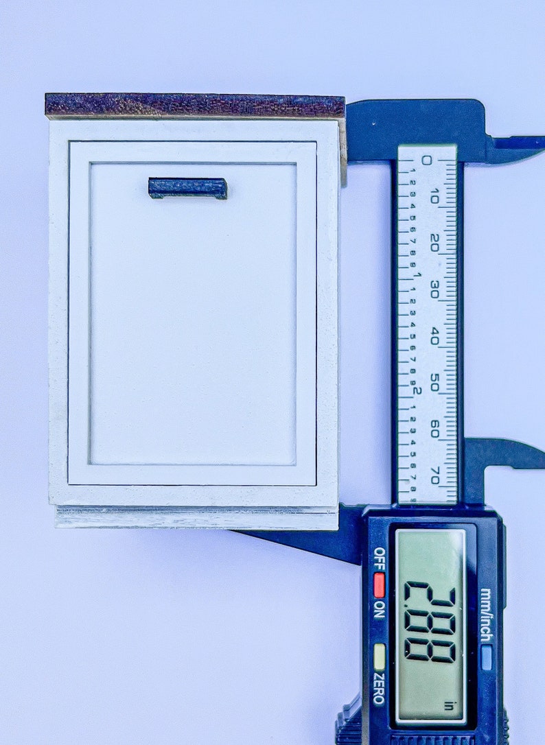 Puede incluir: Un armario blanco con una tapa de madera y una manija azul. Un calibrador digital mide el armario a 7,32 cm.