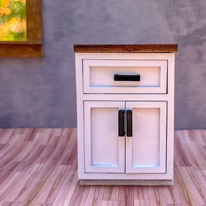 Puede incluir: Un armario blanco con una tapa de madera, un caj&oacute;n y dos puertas. El armario tiene tiradores negros y est&aacute; sobre un suelo de madera.