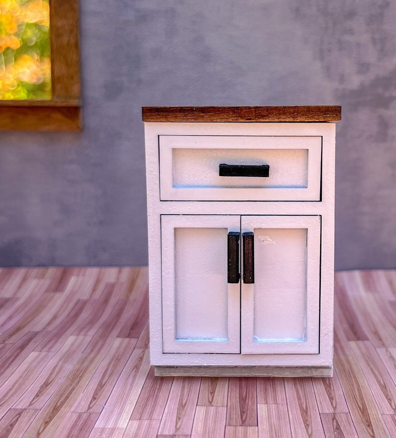 Puede incluir: Un armario blanco con una tapa de madera, un caj&oacute;n y dos puertas. El armario tiene tiradores negros y est&aacute; sobre un suelo de madera.
