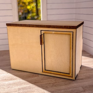 Może przedstawiać: Mały drewniany szafka z jednymi drzwiami. Szafka jest wykonana z jasnego drewna z brązową ramką. Drzwi mają prosty uchwyt i są zamknięte.