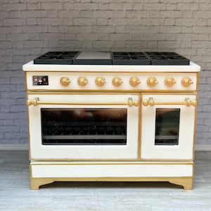 Puede incluir: Un horno en miniatura blanco y dorado con una encimera negra y puertas de horno. El horno tiene perillas y manijas doradas, y una pequeña pantalla digital. Es un electrodoméstico de cocina para casa de muñecas o en miniatura.