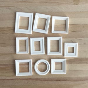 Puede incluir: Una colección de pequeños marcos de fotos blancos de varias formas, incluyendo cuadrados, rectangulares y circulares. Los marcos están dispuestos sobre una superficie de madera marrón claro, creando una exhibición visualmente atractiva para la decoración del hogar o proyectos de manualidades.