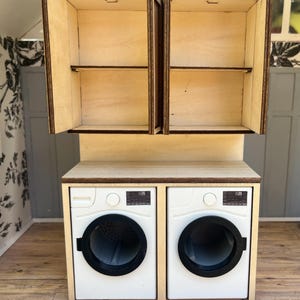 May include: Miniature wooden laundry room set with a pair of white washing machines and a matching cabinet with open doors. The cabinet has two shelves. The set is made of light-colored wood with dark brown trim.