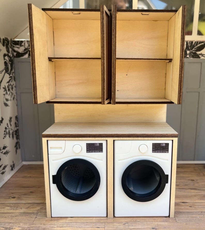May include: Miniature wooden laundry room set with a pair of white washing machines and a matching cabinet with open doors. The cabinet has two shelves. The set is made of light-colored wood with dark brown trim.
