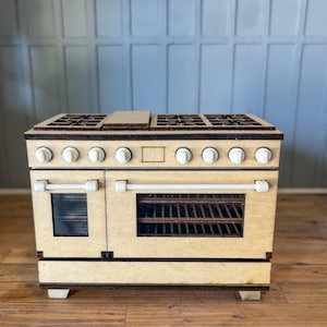 May include: A miniature wooden stove with a light wood finish. The stove features six white knobs, two oven doors with handles, and a cooktop with burners. The oven has a rack inside. The stove is a dollhouse accessory.