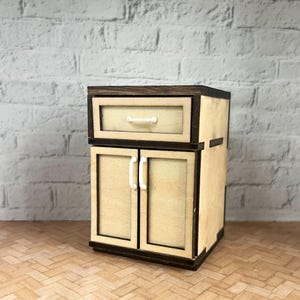 Op de afbeelding: Een kleine houten kast met een lade en twee deuren. De kast is licht van kleur met donkerbruine randen en witte handgrepen. De kast is ontworpen om eruit te zien als een keukenkast.