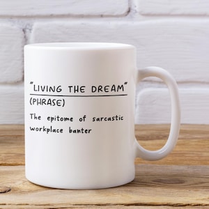 May include: White ceramic mug with a curved handle. The mug features the phrase "LIVING THE DREAM" above the text "(PHRASE)". Below, it reads "The epitome of sarcastic workplace banter" in black text. The mug is on a wooden surface.
