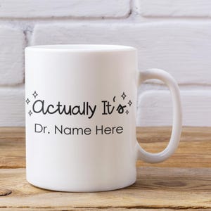 May include: White ceramic mug with a curved handle. The mug features the text "Actually It's" in a handwritten style, followed by "Dr. Name Here" in a simple font. Decorative starbursts are on either side of the first line.