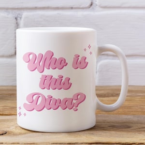 May include: White ceramic mug featuring the text "Who is this Diva?" in pink, retro lettering. The mug has a curved handle and star accents. The mug is on a wooden surface.