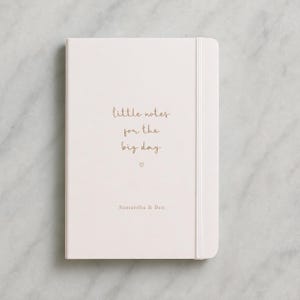 May include: A cream-coloured notebook with the text "little notes for the big day" in gold script. Below the text is a small heart symbol, and the names "Samantha & Ben" are printed at the bottom. The notebook has an elastic band closure.