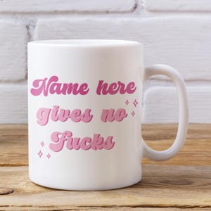 May include: White ceramic mug with a curved handle. The mug features the text "Name here gives no Fucks" in pink, retro-style lettering, with small star accents. The mug is set on a wooden surface.