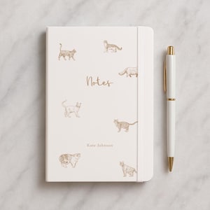 May include: A cream-coloured notebook with a gold pen. The notebook features gold illustrations of cats and the word "Notes" in gold script. The name "Kate Johnson" is printed below the word "Notes". The pen is white with gold accents.