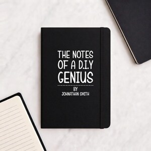May include: Black notebook featuring the white text "THE NOTES OF A D.I.Y GENIUS" and "BY JOHNATHAN SMITH". The notebook has a black elastic band and is next to other notebooks.