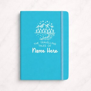 May include: A turquoise notebook featuring a mountain and forest design, with the text "The Travelling Tales of Name Here." It includes an elastic band closure.