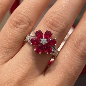 Peut inclure: Une bague argentée avec une pièce centrale en forme de fleur composée de six rubis rouge foncé taille poire entourant un diamant blanc rond central. L'anneau est divisé et incrusté de petits diamants blancs pavés sur les côtés.