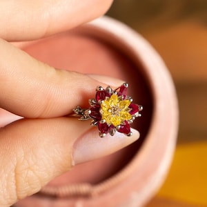 May include: A silver-toned ring featuring a floral design with a cluster of bright yellow marquise-cut stones at the centre, surrounded by a layer of deep red marquise-cut stones, all set in a metallic frame with small clear accent stones.