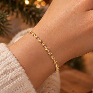 May include: A delicate silver chain bracelet featuring a series of small, round, yellow gemstones set in a row. The bracelet is worn on a wrist, showcasing the bright, circular stones linked together by a thin, polished metallic band.