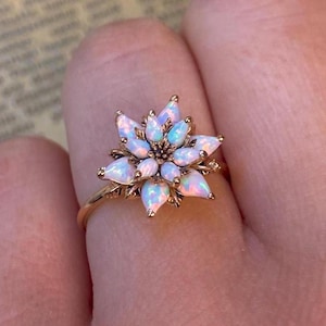 May include: A gold-colored ring featuring a flower design with multiple opal gemstones. The ring's flower has a central cluster of opals surrounded by petal-shaped opals. The ring is on a person's finger, with a book in the background.