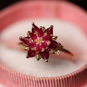May include: A gold ring featuring a central flower-shaped cluster of marquise-cut deep red gemstones. The petals are arranged in layers with a small, clear accent stone on the side of the band, set within a round, light pink velvet ring box.