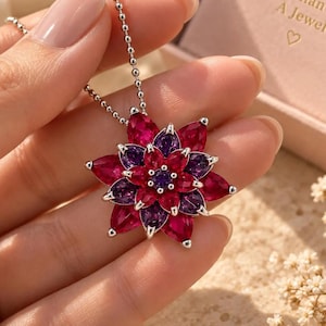 May include: A silver-toned necklace featuring a multi-layered flower pendant with marquise-cut red and purple gemstones. The pendant is suspended from a delicate ball chain. A light pink box is visible in the background with the text More Than A Jewel.