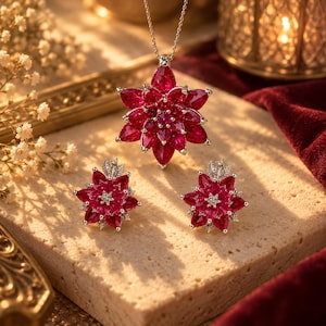 Op de afbeelding: Een sieradenset bestaande uit een ketting met hanger en bijpassende oorbellen, elk met een bloemmotief gemaakt van meerdere dieprode, peervormige edelstenen in lagen, met kleine heldere steentjes in het midden van elk sieraad.