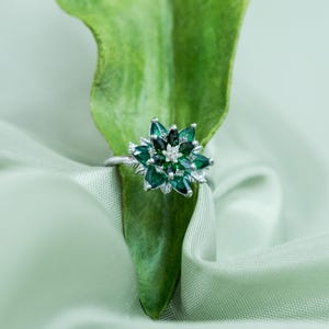 May include: A silver ring with a flower-shaped design. The ring features dark green gemstones arranged in a floral pattern, with a small cluster of white stones at the center. The ring is set against a green leaf and fabric.