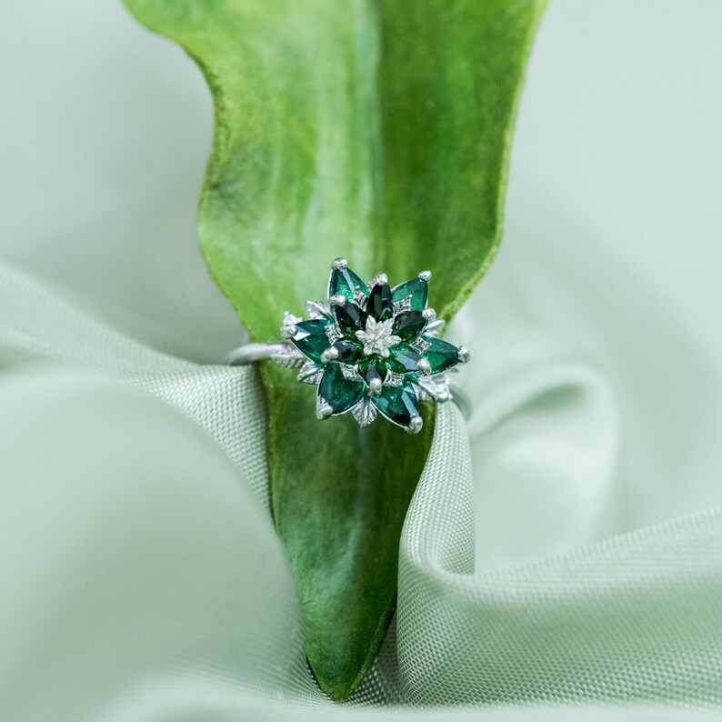 May include: A silver ring with a flower-shaped design. The ring features dark green gemstones arranged in a floral pattern, with a small cluster of white stones at the center. The ring is set against a green leaf and fabric.