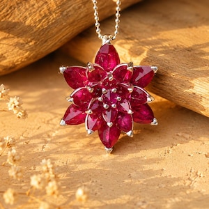 May include: A silver pendant necklace featuring a cluster of deep red, pear-shaped rubies arranged in a floral pattern. The stones are set in a metallic frame with a delicate silver ball chain, creating a symmetrical, multi-layered bloom design.